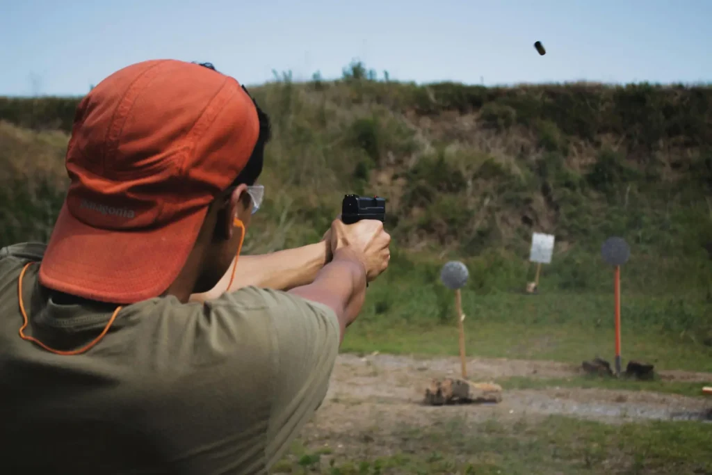 Person in an orange hat firing a handgun at an outdoor shooting range with targets in the background