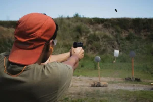 Person in an orange hat firing a handgun at an outdoor shooting range with targets in the background